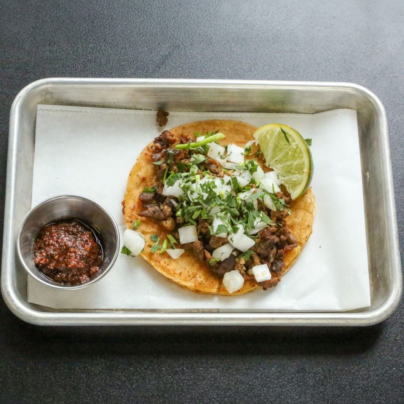 A single taco with meat, cilantro, and diced onions, served with a lime wedge and salsa.