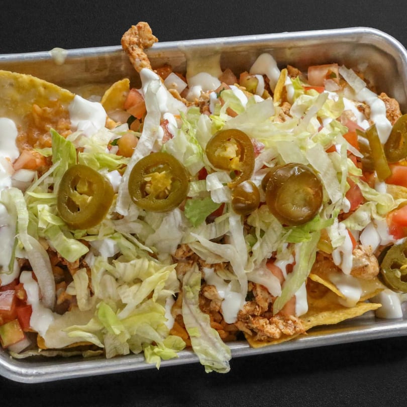 A metal tray filled with nachos topped with chicken, lettuce, tomatoes, onions, sour cream, and jalapeños.