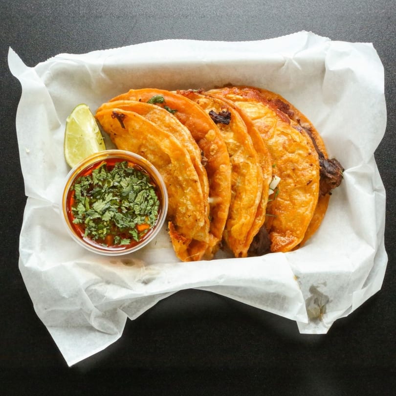 Five crispy birria tacos with a side of consommé and a lime wedge in a white paper-lined basket.